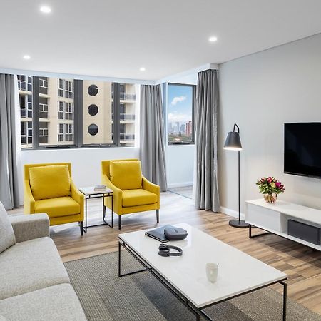 Meriton Suites Bondi Junction Sídney Exterior foto The photo shows a modern, well-lit living room. It features a light gray sofa with a textured fabric. There are two bright yellow armchairs positioned near a window, which allows natural light to enter the space. The room has light gray curtains and 
