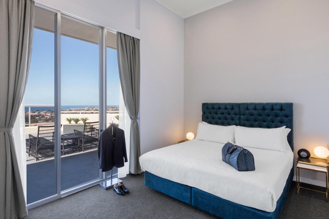 Meriton Suites Bondi Junction Sídney Exterior foto The photo shows a stylish bedroom featuring a large bed with a tufted blue headboard. There's a neatly organized outfit consisting of a suit hanging on a stand, along with shoes placed on the floor. The room has large windows that provide a view of t