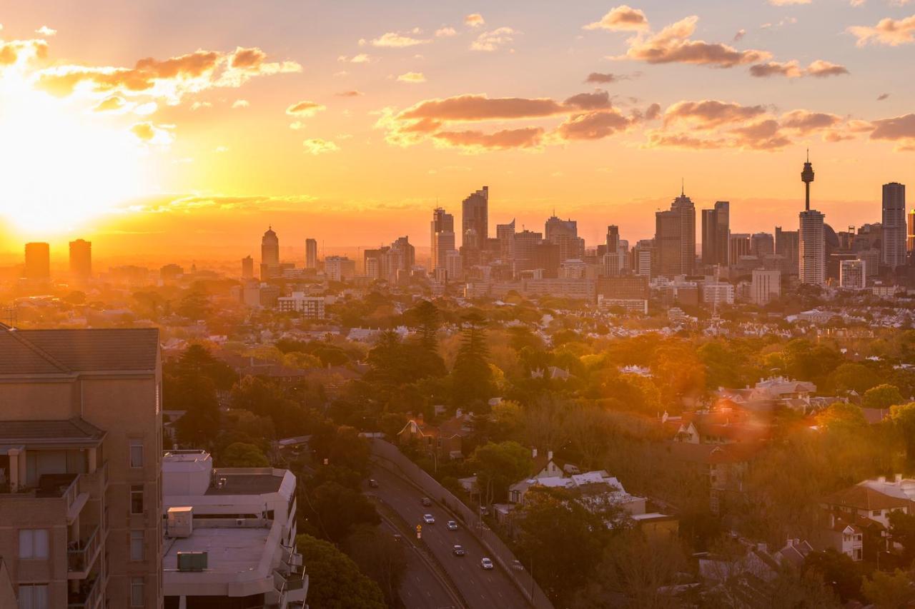 Meriton Suites Bondi Junction Sídney Exterior foto The photo shows a panoramic view of a city skyline at sunset. The sun is low on the horizon, casting a warm golden light across the buildings. Tall skyscrapers are silhouetted against the colorful sky, which features hues of orange, pink, and yellow,