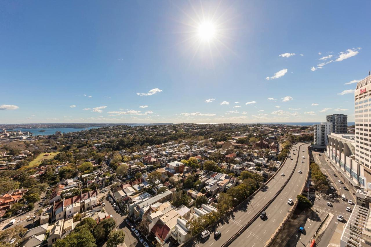 Meriton Suites Bondi Junction Sídney Exterior foto The photo shows a panoramic view of a city landscape under a clear blue sky. In the foreground, there is a road with vehicles traveling along it, bordered by trees and buildings. In the background, you can see a residential area with various houses, 