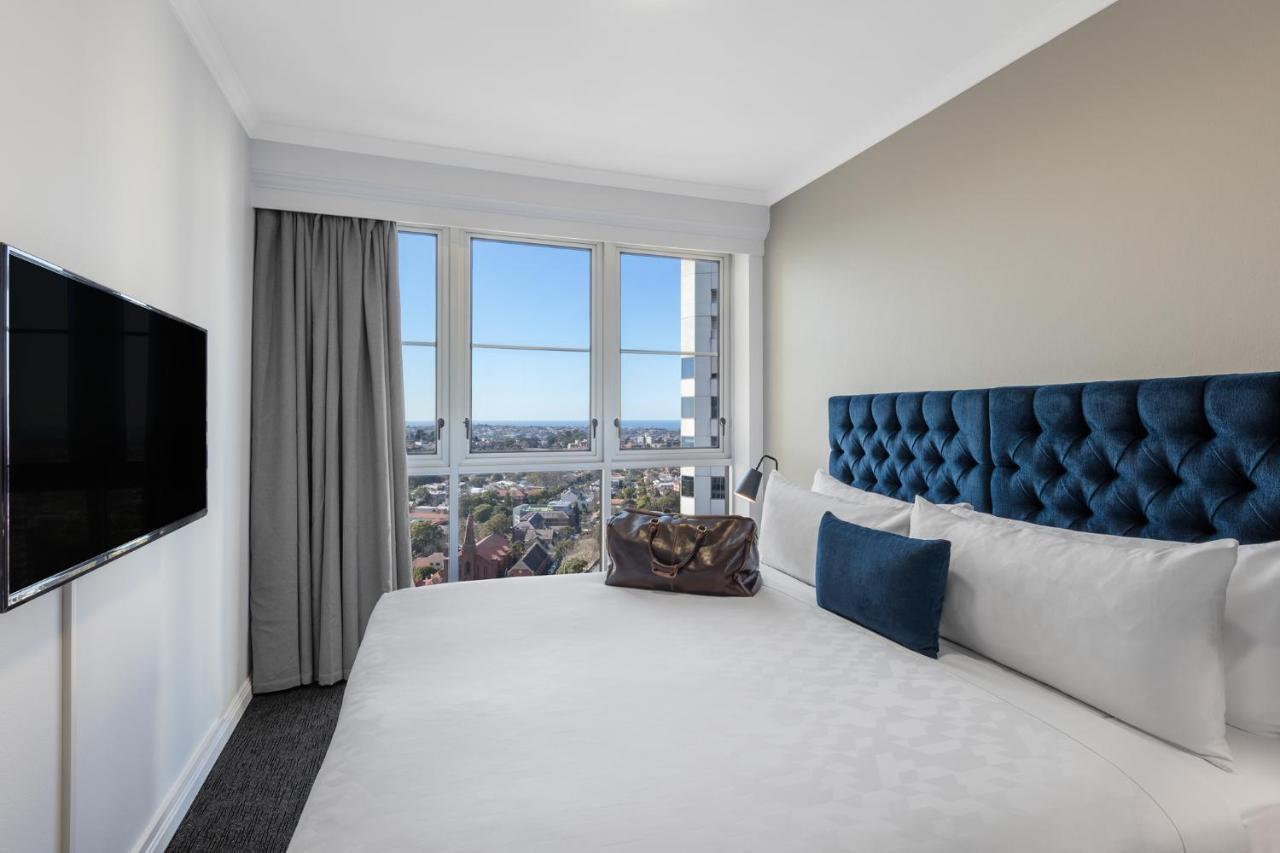 Meriton Suites Bondi Junction Sídney Exterior foto The photo shows a modern hotel room featuring a large bed with a tufted blue headboard. Beside the bed, there is a stylish bag placed on the bed. The room has large windows that provide a view of the outside, allowing natural light to fill the space.