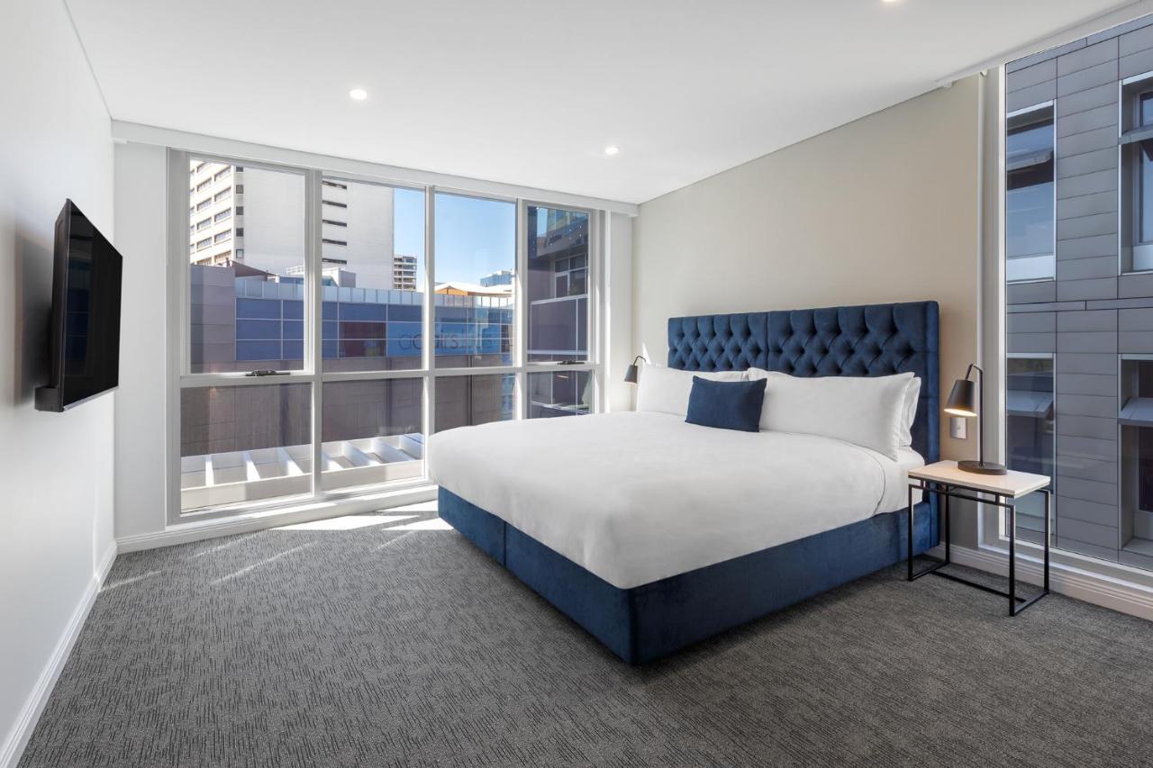 Meriton Suites Bondi Junction Sídney Exterior foto The image shows a modern hotel room with a large, comfortable bed featuring a blue upholstered headboard. The room has large windows that allow natural light to enter, providing a good view of the surrounding buildings. There is a small bedside table