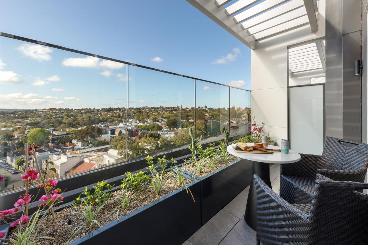 Meriton Suites Bondi Junction Sídney Exterior foto The photo shows a modern balcony with a glass railing, enhancing the view of the surrounding area, which includes various rooftops and greenery in the distance. There is a small round table set with a decorative floral arrangement and some items, lik