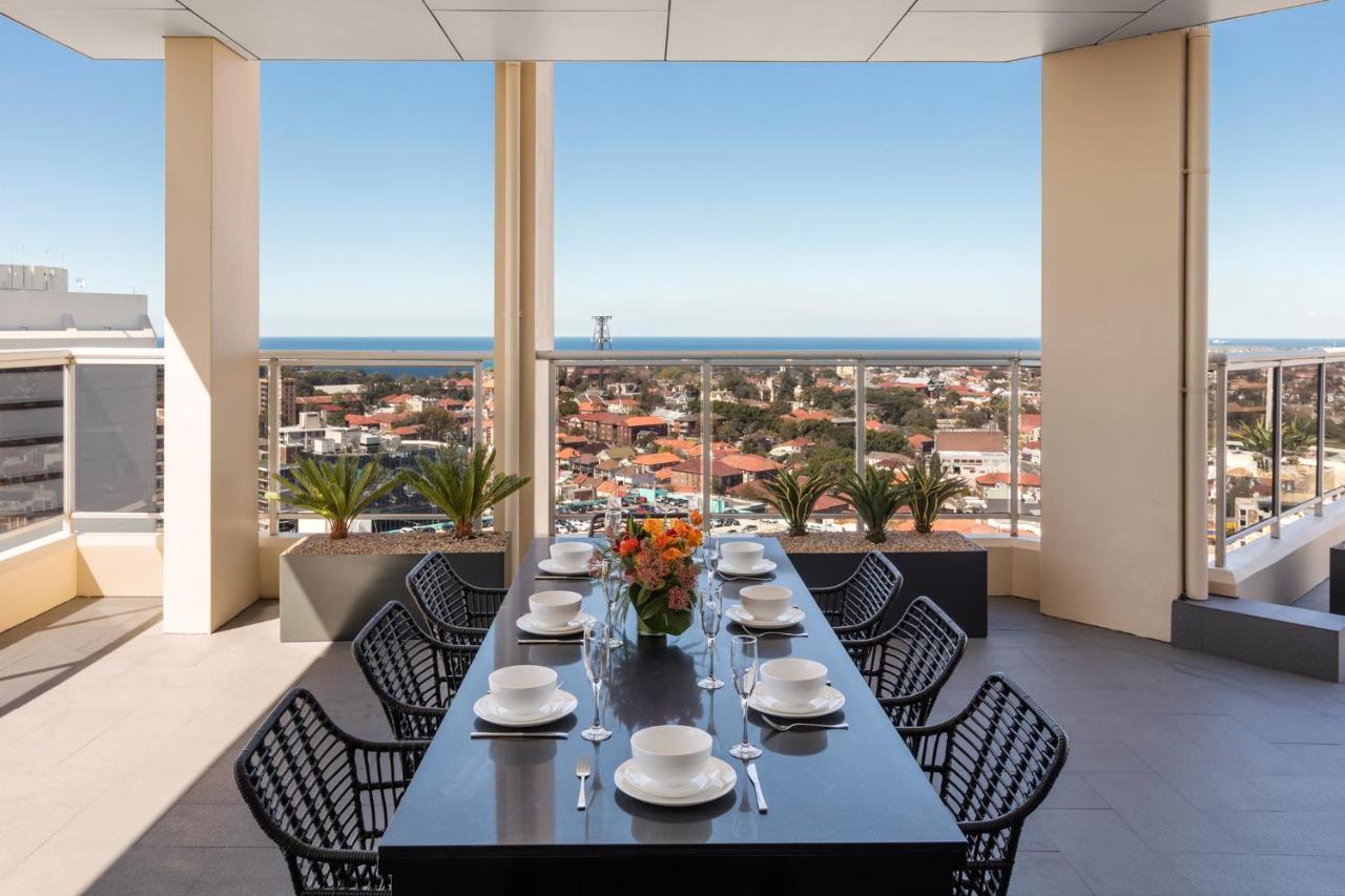Meriton Suites Bondi Junction Sídney Exterior foto The photo shows a modern outdoor dining area situated on a balcony or terrace with a stunning view. There is a long rectangular table set with plates, glasses, and cutlery, ready for a meal. The table is surrounded by sleek, black wicker chairs. In t