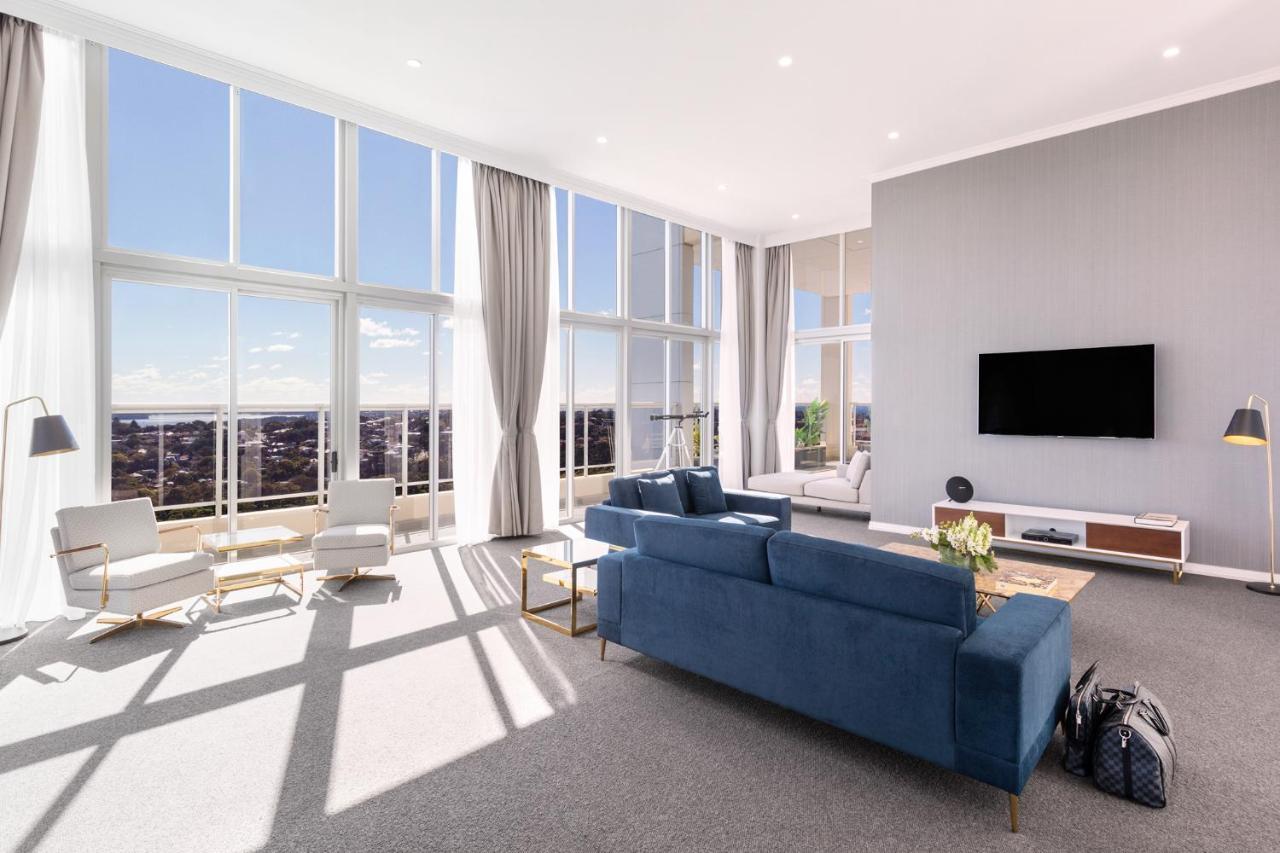 Meriton Suites Bondi Junction Sídney Exterior foto The photo shows a bright and modern living room with large floor-to-ceiling windows that provide a view of the outdoors. The space features a blue sofa in the center, complemented by light-colored armchairs nearby. There are light curtains hanging by
