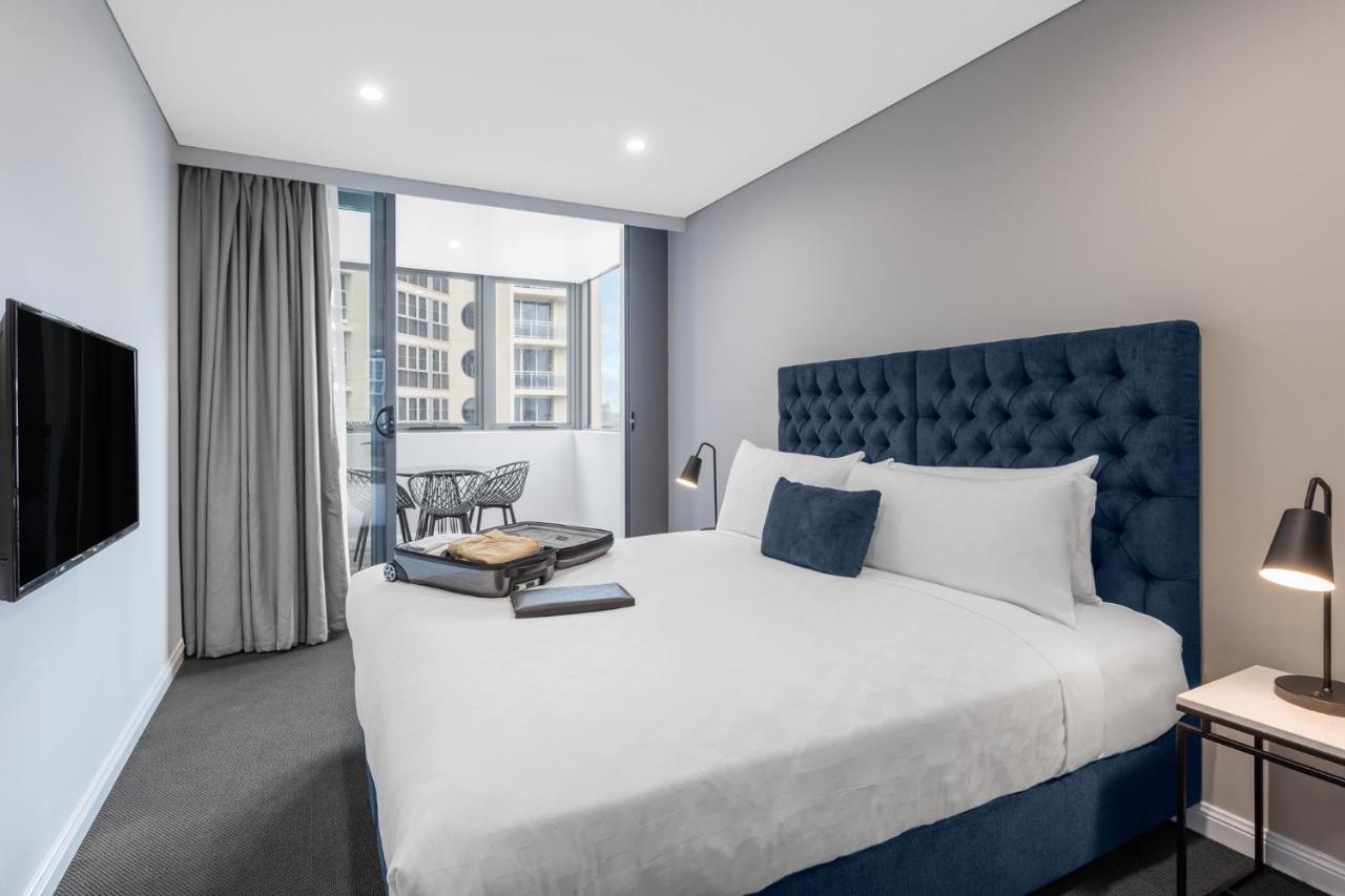 Meriton Suites Bondi Junction Sídney Exterior foto The photo shows a modern hotel room featuring a large bed with a tufted blue headboard. There are minimalistic bedside tables with lamps on either side of the bed. A flat-screen TV is mounted on the wall across from the bed. To one side, there are fl
