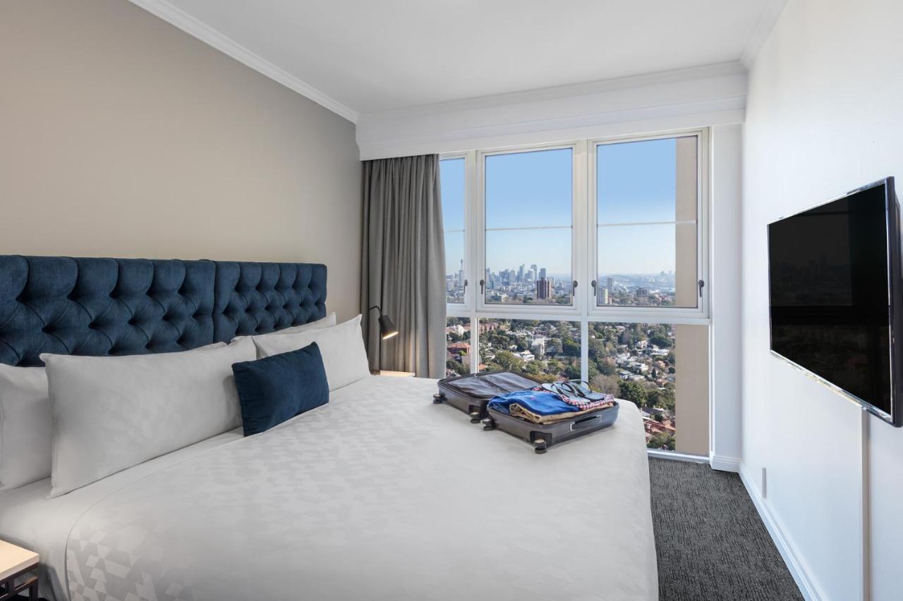 Meriton Suites Bondi Junction Sídney Exterior foto The photo shows a modern hotel bedroom featuring a king-sized bed with a blue tufted headboard. The bed is neatly made with white linens and a decorative blue pillow. There is a large window that offers a view of the city skyline, allowing natural li