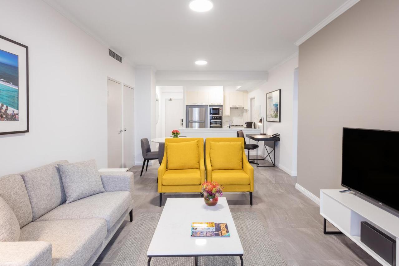 Meriton Suites Bondi Junction Sídney Exterior foto The photo shows a modern living space with a light and airy design. In the foreground, there are two bright yellow armchairs flanking a white coffee table, which has a small vase of flowers and a magazine on it. To the left, there is a light gray sof