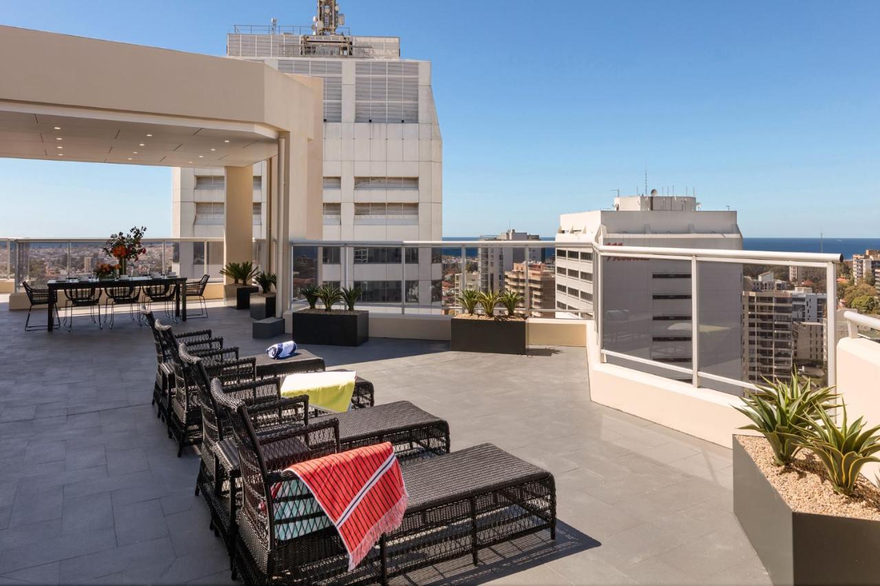 Meriton Suites Bondi Junction Sídney Exterior foto The photo shows a rooftop terrace with a modern design. There are several outdoor seating arrangements featuring black wicker lounge chairs. Some of the chairs have colorful blankets draped over them. The terrace is surrounded by a railing and has po
