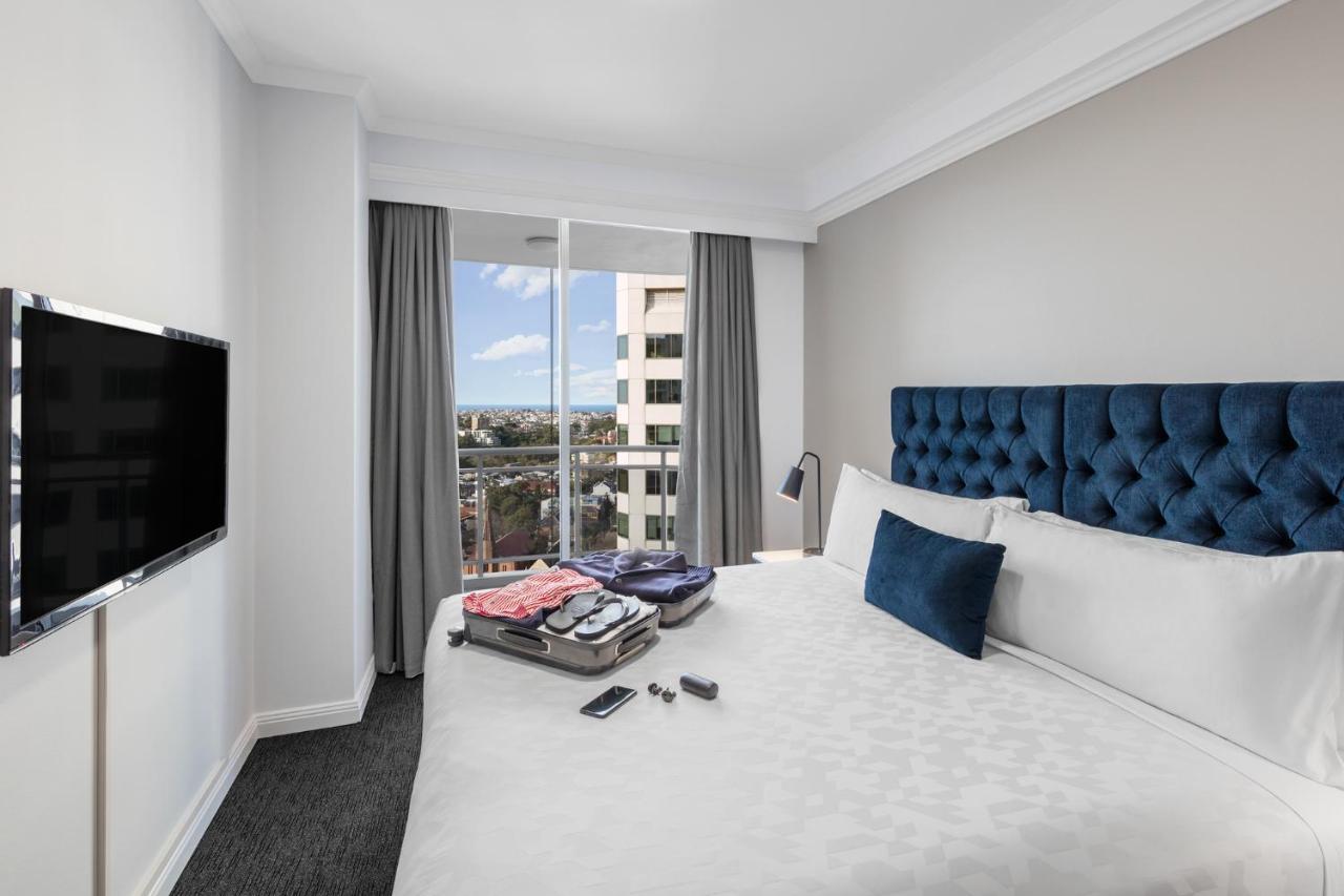 Meriton Suites Bondi Junction Sídney Exterior foto The photo shows a modern hotel room featuring a neatly made bed with a blue tufted headboard and a white duvet. There's a small decorative pillow on the bed, and a piece of luggage is placed at the foot with clothes partially unpacked. A television i