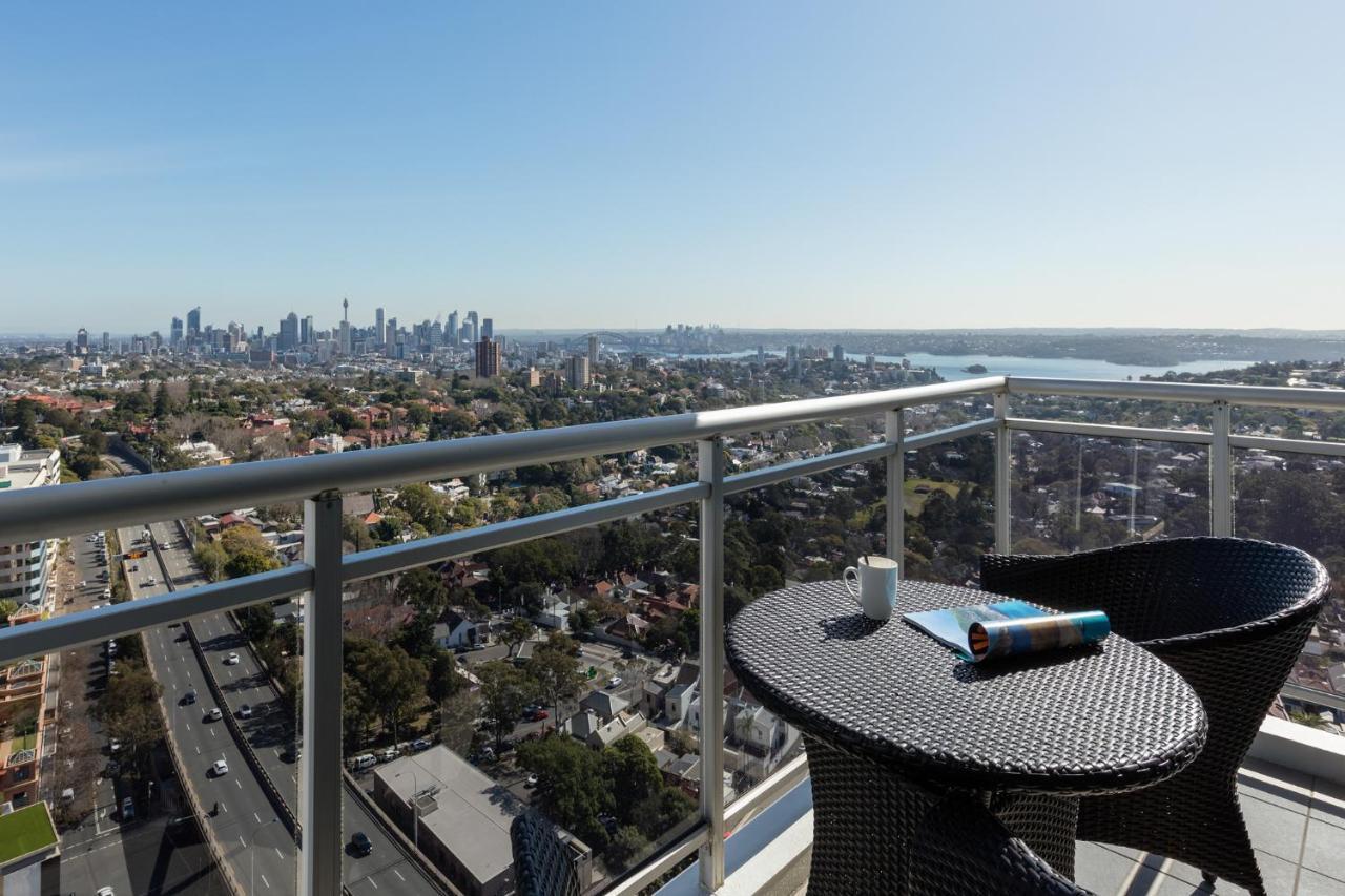 Meriton Suites Bondi Junction Sídney Exterior foto The photo shows a balcony with a view overlooking a cityscape and a body of water in the distance. There is a round table with a woven design and two chairs, one of which has a mug on it. On the table, there’s also a rolled-up magazine or document. T