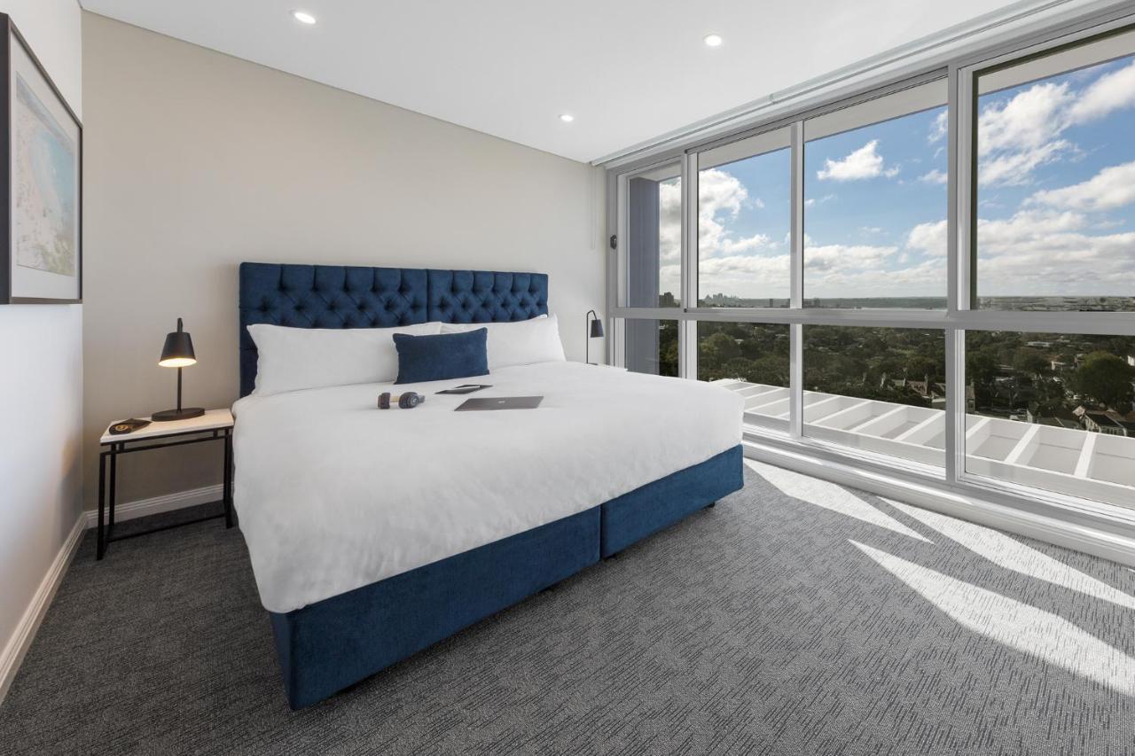 Meriton Suites Bondi Junction Sídney Exterior foto The photo shows a modern and stylish hotel room. It features a comfortable queen-size bed with a blue tufted headboard. The bed is neatly made with white linens and pillows. Next to the bed, there is a small bedside table with a desk lamp. 

On the f
