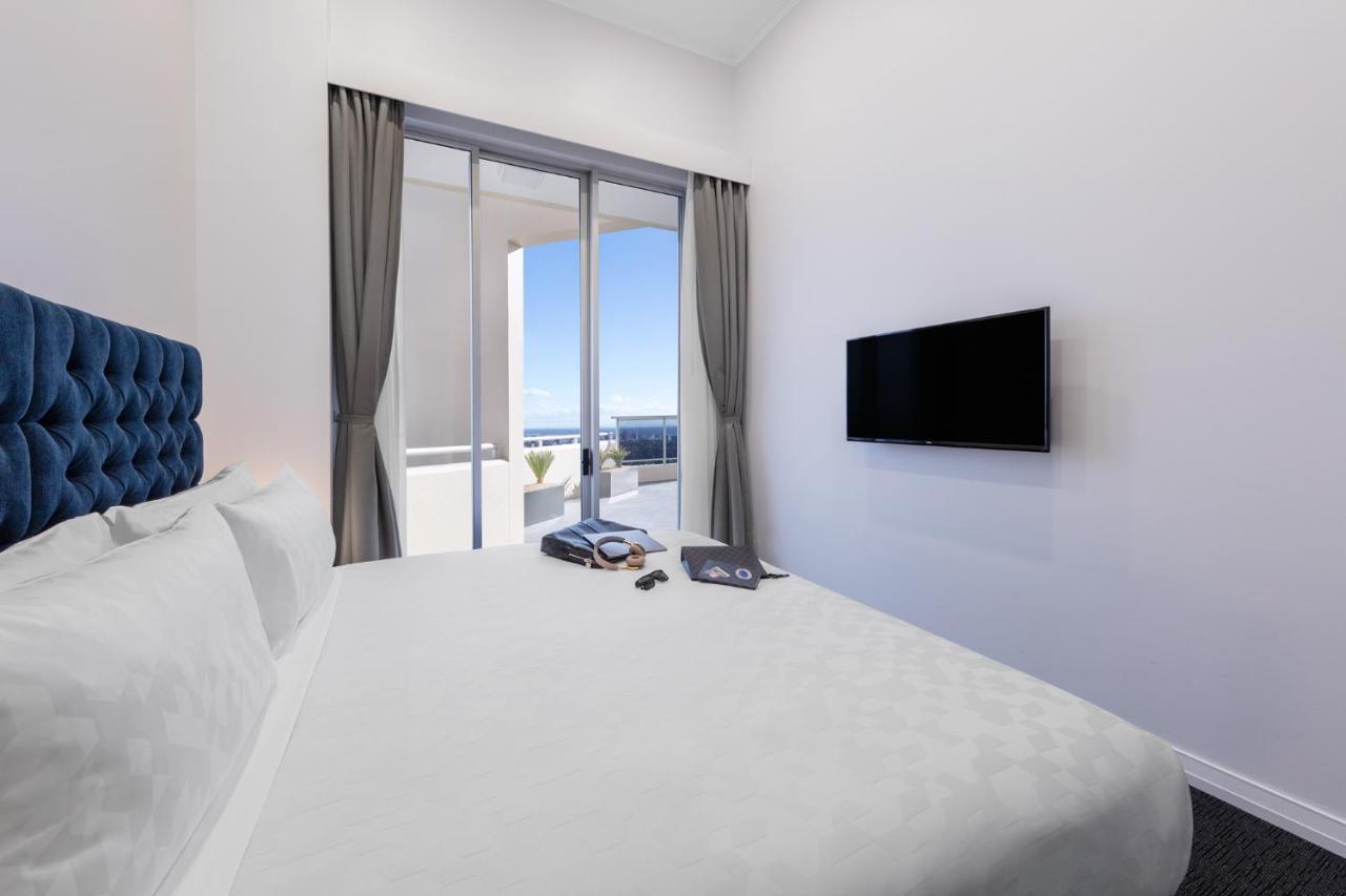 Meriton Suites Bondi Junction Sídney Exterior foto The photo shows a modern hotel room with a large, comfortable bed featuring a blue tufted headboard. There are light-colored walls, and the room is decorated simply. A flat-screen TV is mounted on the wall opposite the bed. The room has large glass d