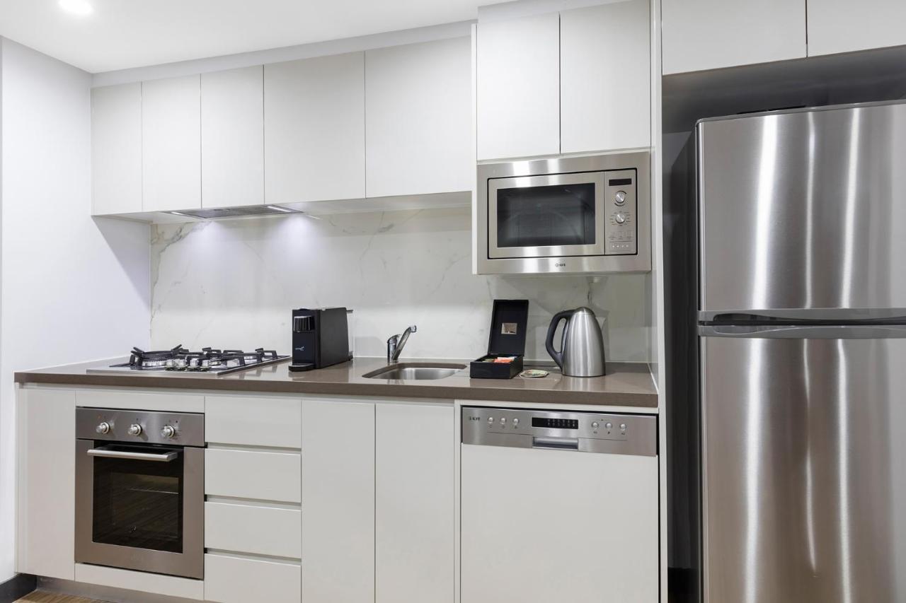 Meriton Suites Bondi Junction Sídney Exterior foto The photo shows a modern kitchen with a sleek, minimalist design. It features white cabinetry and a countertop that blends with the overall aesthetic. 

Key elements include:

- A stainless steel refrigerator on the right.
- An oven with a gas cookto
