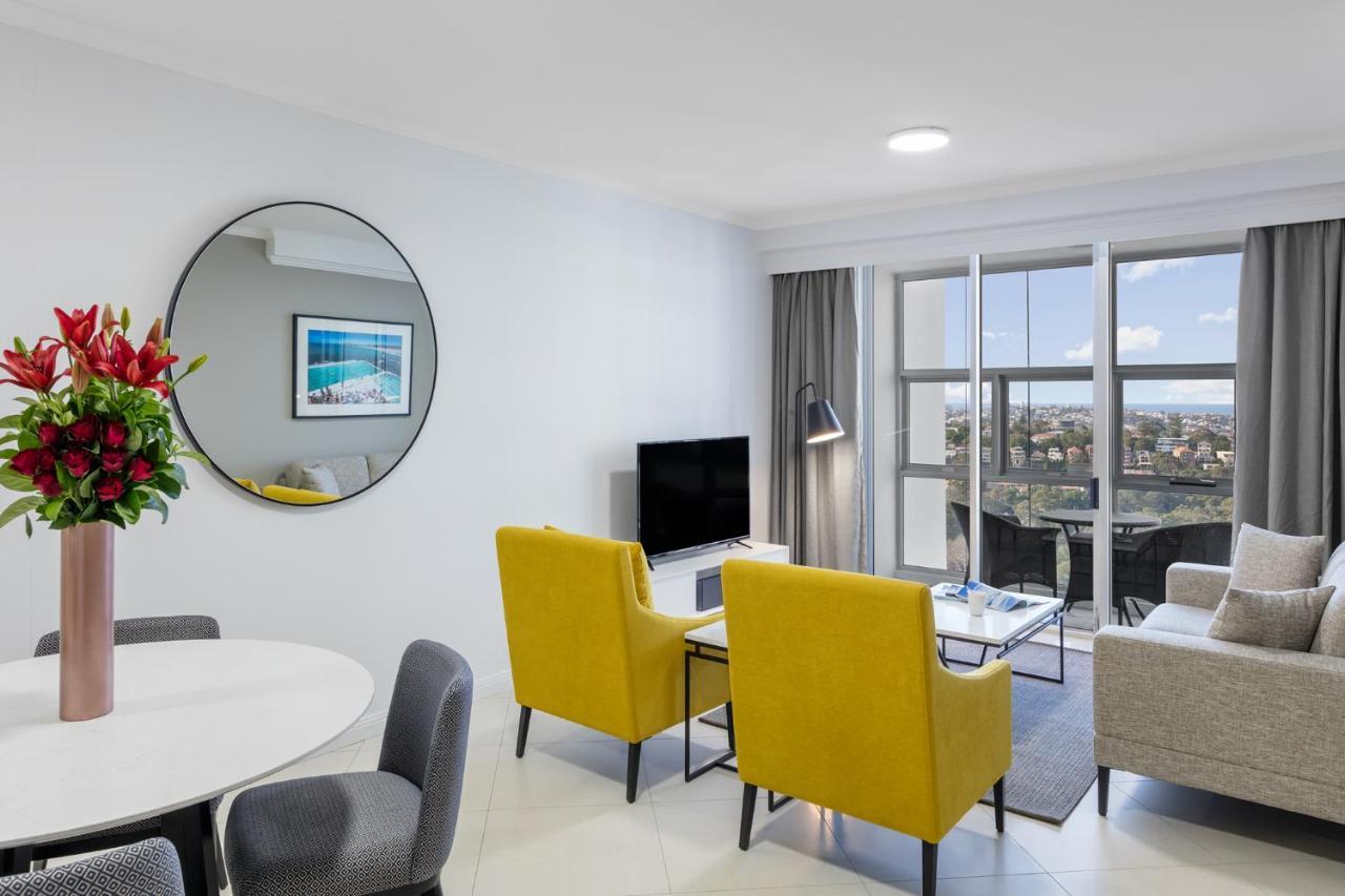 Meriton Suites Bondi Junction Sídney Exterior foto The photo shows a modern living space with a stylish design. There are two bright yellow chairs facing a small glass table, and a comfortable beige sofa to the right. A TV is positioned on the wall in front of the chairs. To the left, there's a dinin