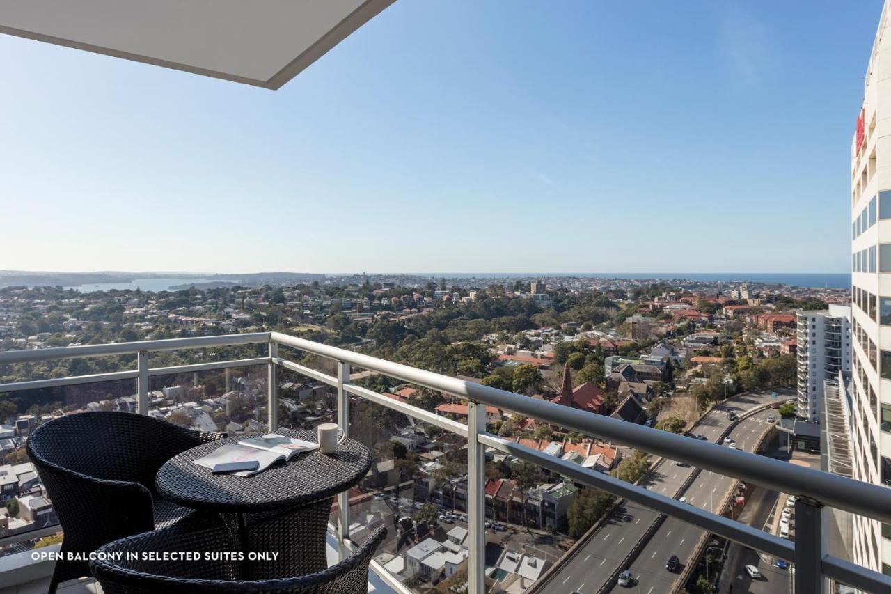 Meriton Suites Bondi Junction Sídney Exterior foto The photo shows a balcony with a view over a cityscape that includes buildings and greenery, extending towards the horizon where the sea is visible. The balcony features a small round table and two black chairs, suggesting a space for relaxation or d