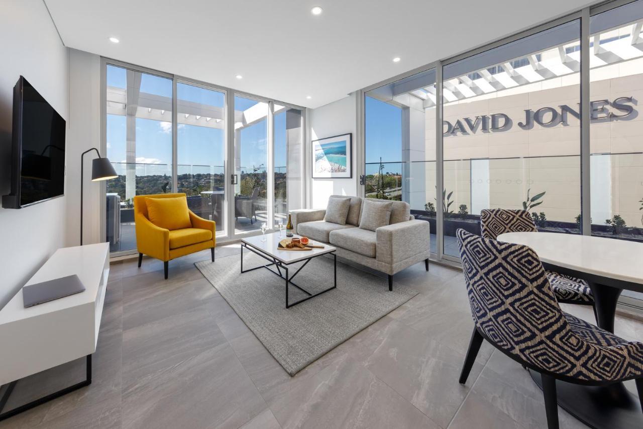 Meriton Suites Bondi Junction Sídney Exterior foto The photo shows a modern living space that features large windows allowing ample natural light. The room is furnished with a gray sofa and a bright yellow armchair, along with a coffee table in between. There’s a decorative bowl with fruits on the ta