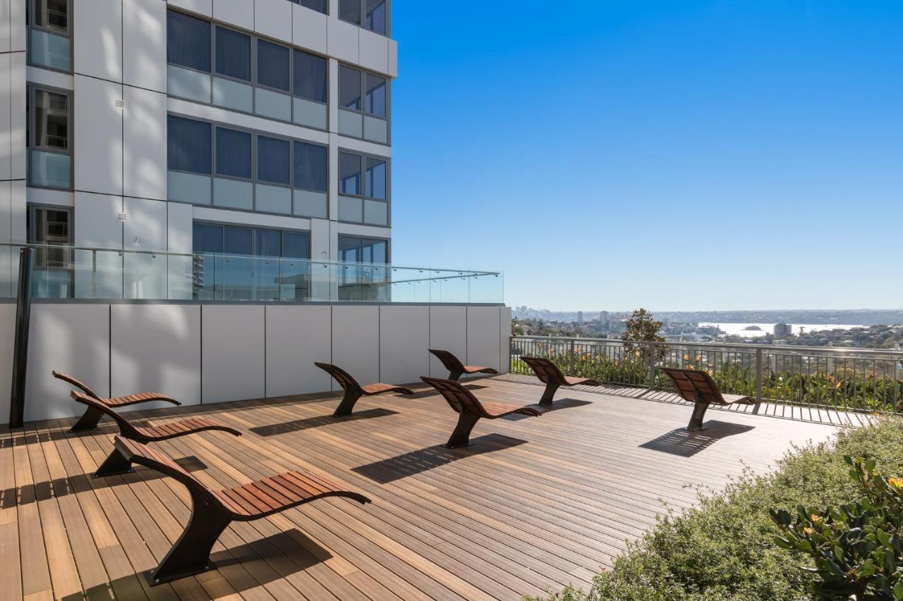 Meriton Suites Bondi Junction Sídney Exterior foto The photo shows a rooftop or outdoor area with a wooden deck. There are several modern lounge chairs arranged neatly, facing a clear sky and a panoramic view of the surrounding landscape. In the background, there is a tall building with large windows