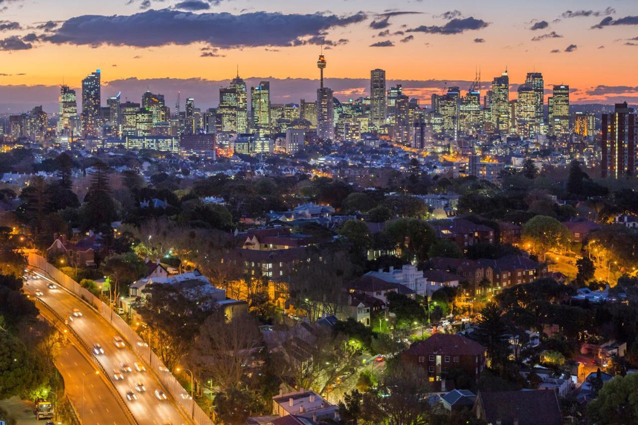 Meriton Suites Bondi Junction Sídney Exterior foto The photo captures a stunning city skyline at dusk, featuring towering skyscrapers adorned with lights against a colorful sunset backdrop. In the foreground, a green area with trees and houses is visible, while a road with cars can be seen winding th