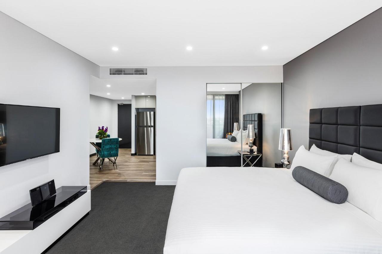 Meriton Suites Bondi Junction Sídney Exterior foto The photo shows a modern hotel room. It features a large bed with a white covering and gray accent pillows. To the left, there's a flat-screen TV mounted on the wall above a sleek white cabinet. The flooring is a light wood tone, and a large mirror i