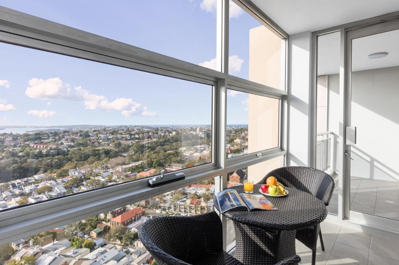 Meriton Suites Bondi Junction Sídney Exterior foto The photo shows a modern balcony with large glass windows that offer a panoramic view of a sprawling urban landscape. There is a small round table made of dark wicker, accompanied by two matching chairs. On the table, there is a bowl of fruit and a m