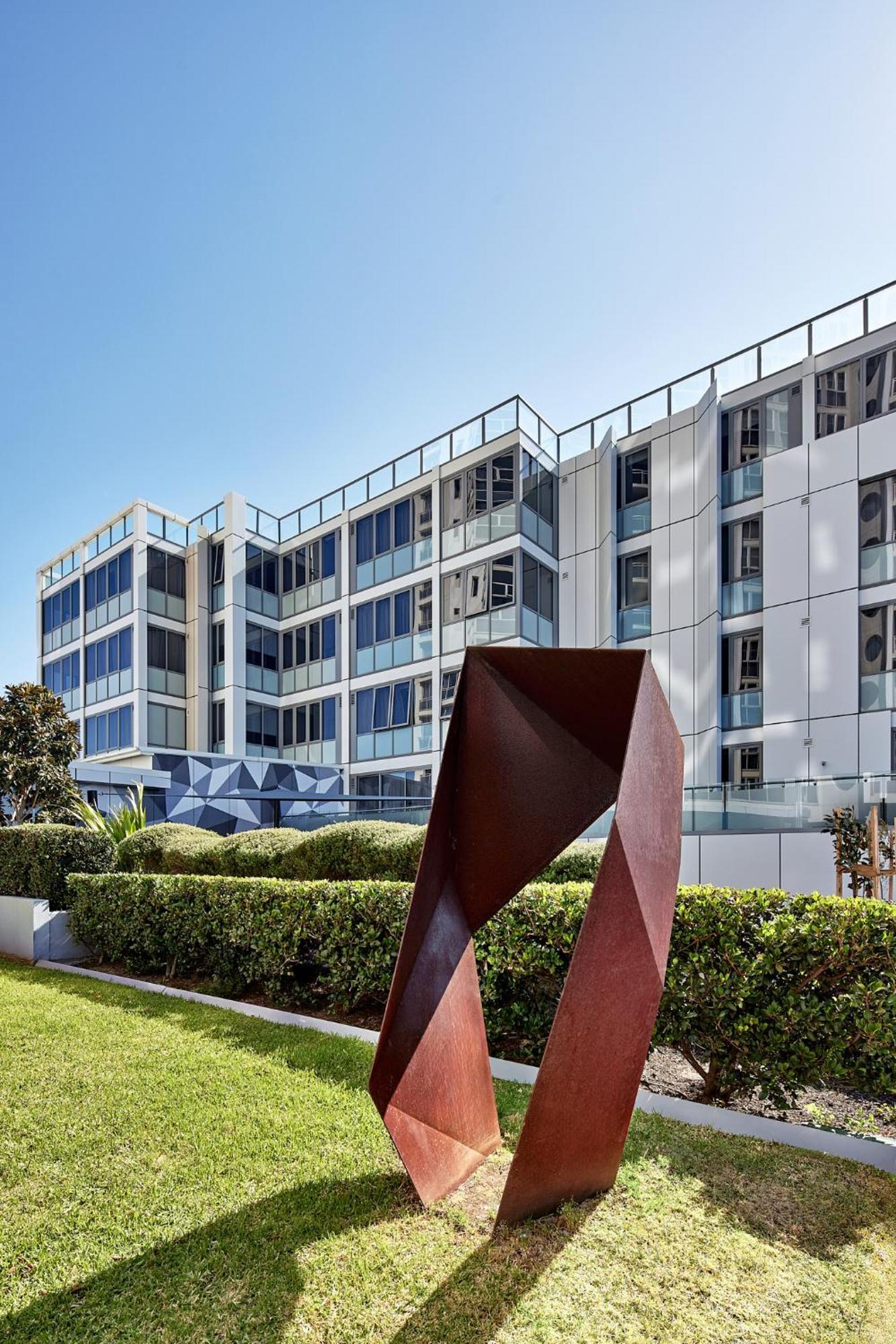 Meriton Suites Bondi Junction Sídney Exterior foto The photo shows a modern building with a sleek white and glass facade, featuring multiple floors and large windows. In the foreground, there is a sculptural piece made of metal, characterized by geometric shapes and a reddish-brown color. The surroun