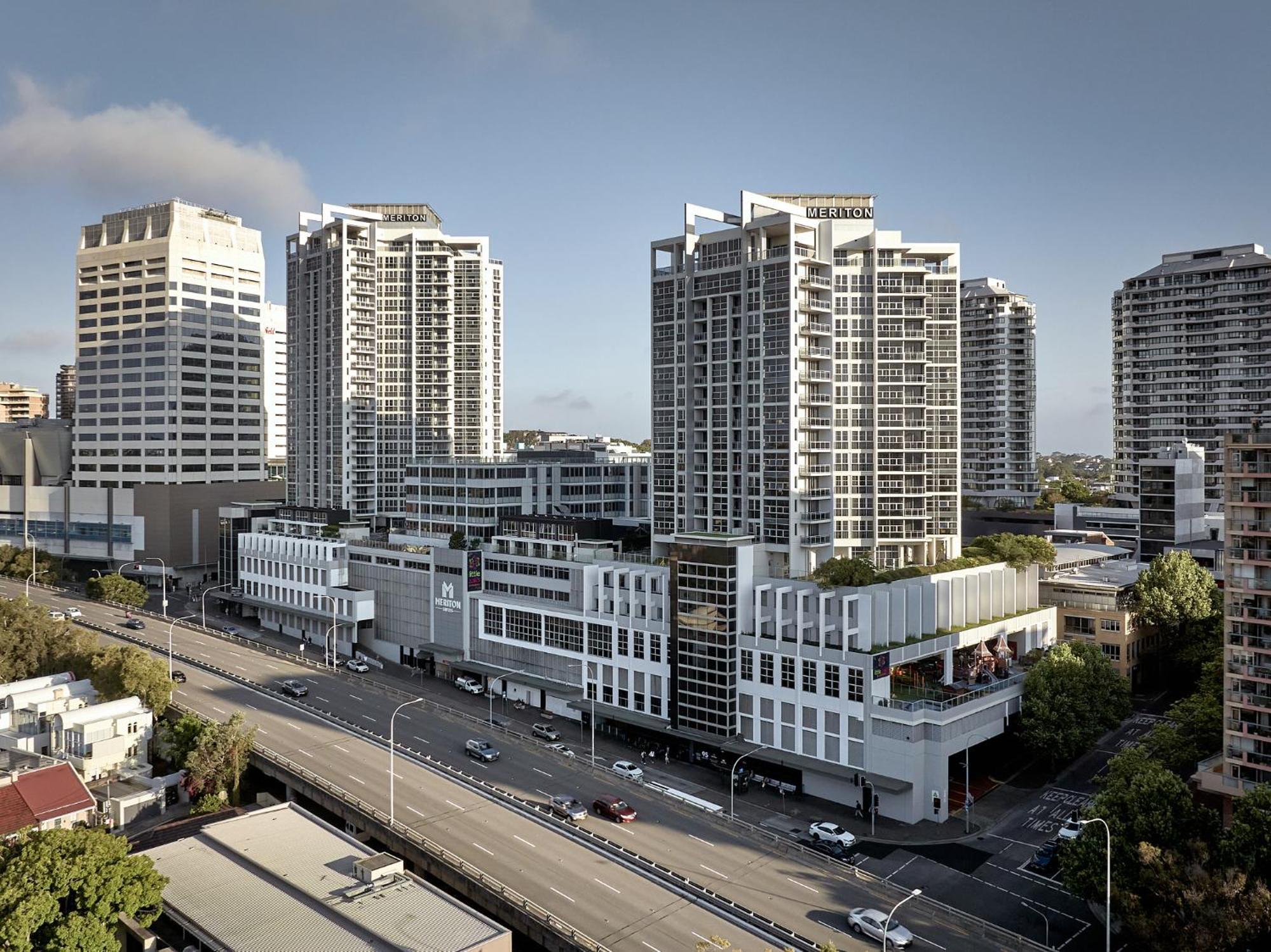 Meriton Suites Bondi Junction Sídney Exterior foto The photo shows a cityscape featuring several tall modern buildings, likely apartments or office complexes. In the foreground, there is a road lined with cars, and the architecture of the buildings varies, with some having sleek glass facades and oth