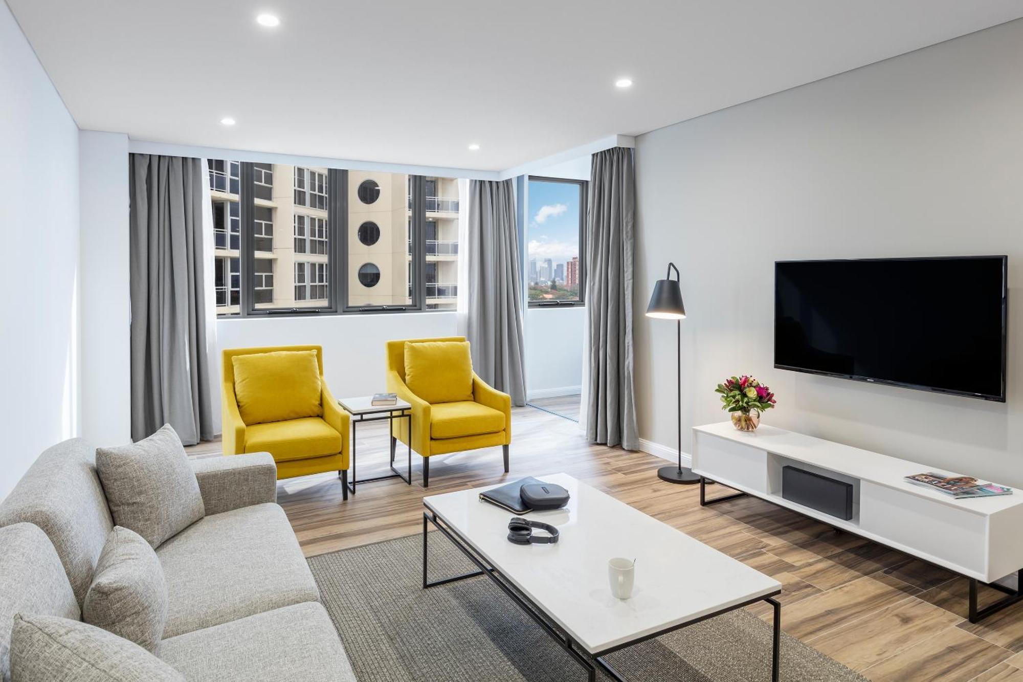 Meriton Suites Bondi Junction Sídney Exterior foto The photo shows a modern, well-lit living room. It features a light gray sofa with a textured fabric. There are two bright yellow armchairs positioned near a window, which allows natural light to enter the space. The room has light gray curtains and 